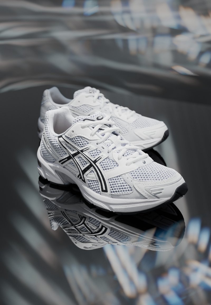 Paire de chaussures de course en mesh blanc avec des accents noirs, présentée sur une surface réfléchissante en intérieur.