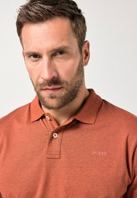 Kortærmet polo skjorte i rust orange, lavet af tekstureret stof. Har en tre-knaps knaplukning og et diskret logo på brystet.