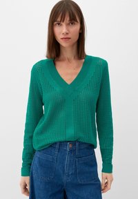Groene gebreide trui met een V-hals, voorzien van geperforeerde patronen en lange mouwen, gecombineerd met hoge taille blauwe spijkerbroek.