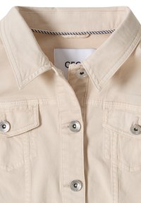 Béžová bunda s límcem se stříbrnými cvoky a dvěma kapsami na prsou se zapínáním na knoflíky, zobrazující detailní šití a vnitřní štítek.