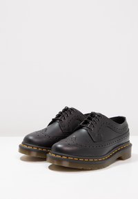 Černé kožené společenské boty mají perforované brogue detaily, kulatou špičku a silnou průhlednou gumovou podrážku s žlutými švy.