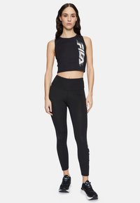 Top corto negro con logo blanco, combinado con leggings negros de cintura alta. La modelo lleva zapatillas deportivas negras con toques azules.