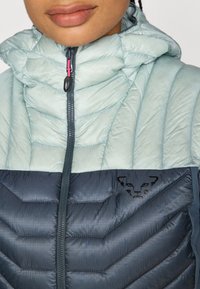 Geacă pufoasă de un albastru deschis cu glugă, având o secțiune inferioară de un albastru închis și un logo negru, confecționată din material texturat și captusit.