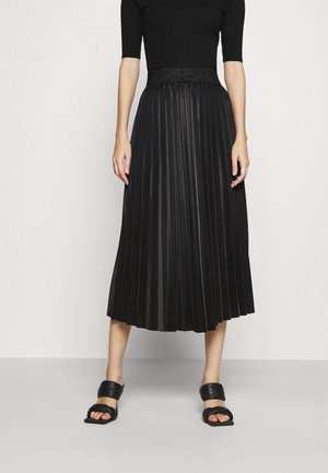 Woman wearing a black pleated midi skirt, black short-sleeve top, and black open-toe sandals with wide straps, standing against a white background.