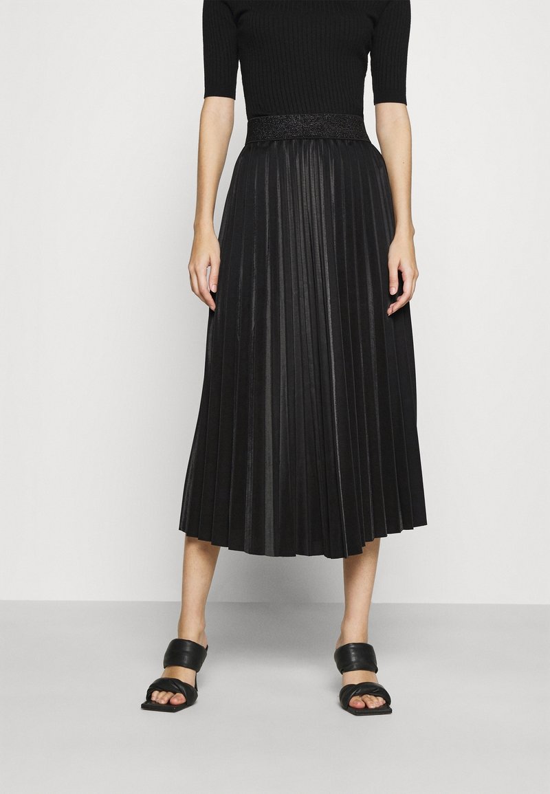 Femme portant une jupe midi plissée noire, un haut noir à manches courtes et des sandales noires à larges brides ouvertes, debout devant un fond blanc.