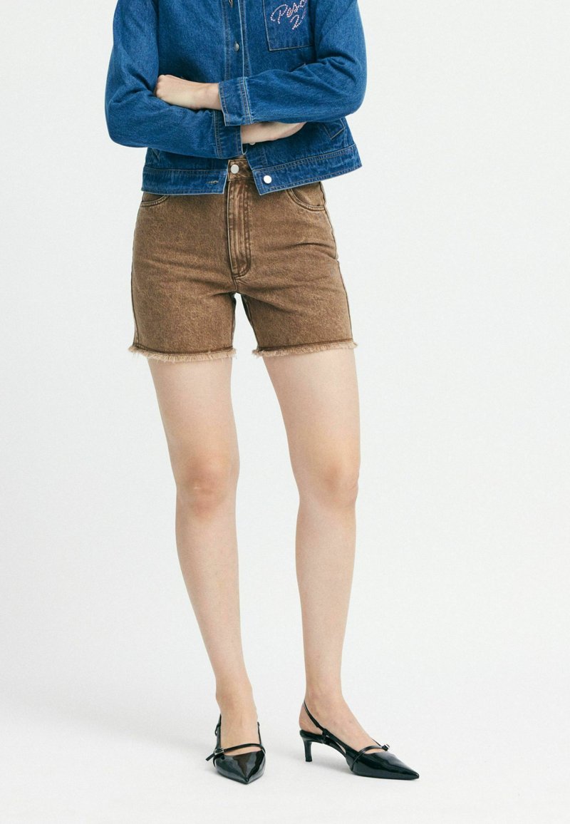 Chaqueta de mezclilla en azul con bolsillo en el pecho y logotipo bordado, combinada con pantalones cortos marrones de tiro alto con dobladillo deshilachado, y tacones negros puntiagudos.