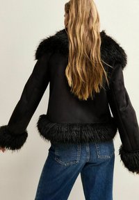 Mujer con pelo largo rubio que lleva una chaqueta negra con borde de piel sintética en el cuello, los puños y el dobladillo, combinada con jeans de mezclilla azul, vista desde atrás.