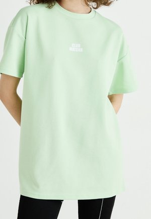 Osoba nosząca jasnooliwkowy oversize t-shirt z napisem "CLUB MATCHA" w białym kolorze na piersi, połączony z czarnymi spodniami.