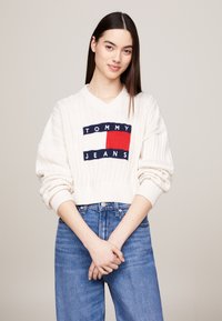 Weißer, gerippter, gestrickter Crop-Pullover mit V-Ausschnitt, ausgestattet mit einem Logo-Patch in Marineblau und Rot. Kombiniert mit hochtaillierten blauen Jeans.