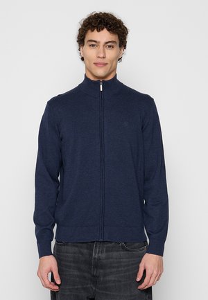 Mann mit lockigem Haar, der einen dunkelblauen Reißverschluss-Pullover und dunkle Jeans trägt und vor einem schlichten hellen Hintergrund steht.