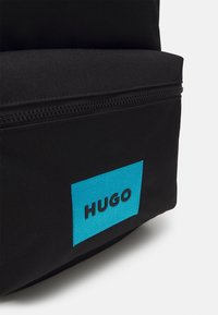 Sac à dos en tissu noir avec un patch rectangulaire bleu proéminent portant le mot "HUGO" en noir. Comprend un compartiment zippé.