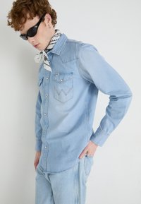 Camisa de mezclilla azul claro con botones a presión, dos bolsillos en el pecho con solapas puntiagudas y costuras en contraste. Combinada con un accesorio de bufanda estampada.
