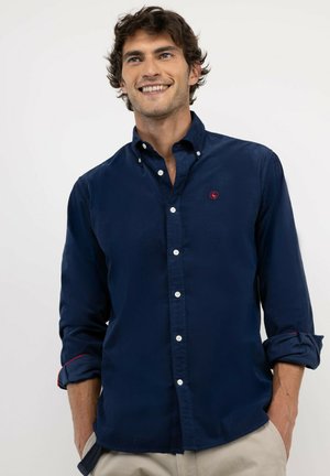 Hombre sonriendo, vestido con una camisa azul marino abotonada con las mangas remangadas y pantalones beige, de pie frente a un fondo blanco liso.