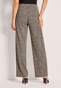 Wijde broek in een visgraatpatroon, met een gestructureerde bruine en zwarte stof, een hoge taille en een platte voorkant. Een paar zwarte hakken zichtbaar.