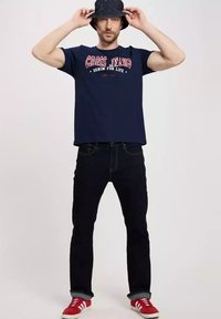 Granatowa koszulka z wyraźnym czerwonym napisem. Połączona z ciemnymi dżinsami i czerwonymi sneakersami. Model nosi czarny kapelusz typu bucket.