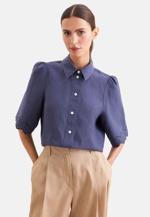 Kvinde med mørkt hår i en lav hestehale iført en blå skjorte med knapper og puffede ærmer samt beige bukser med høj talje, stående med hænderne i lommerne.