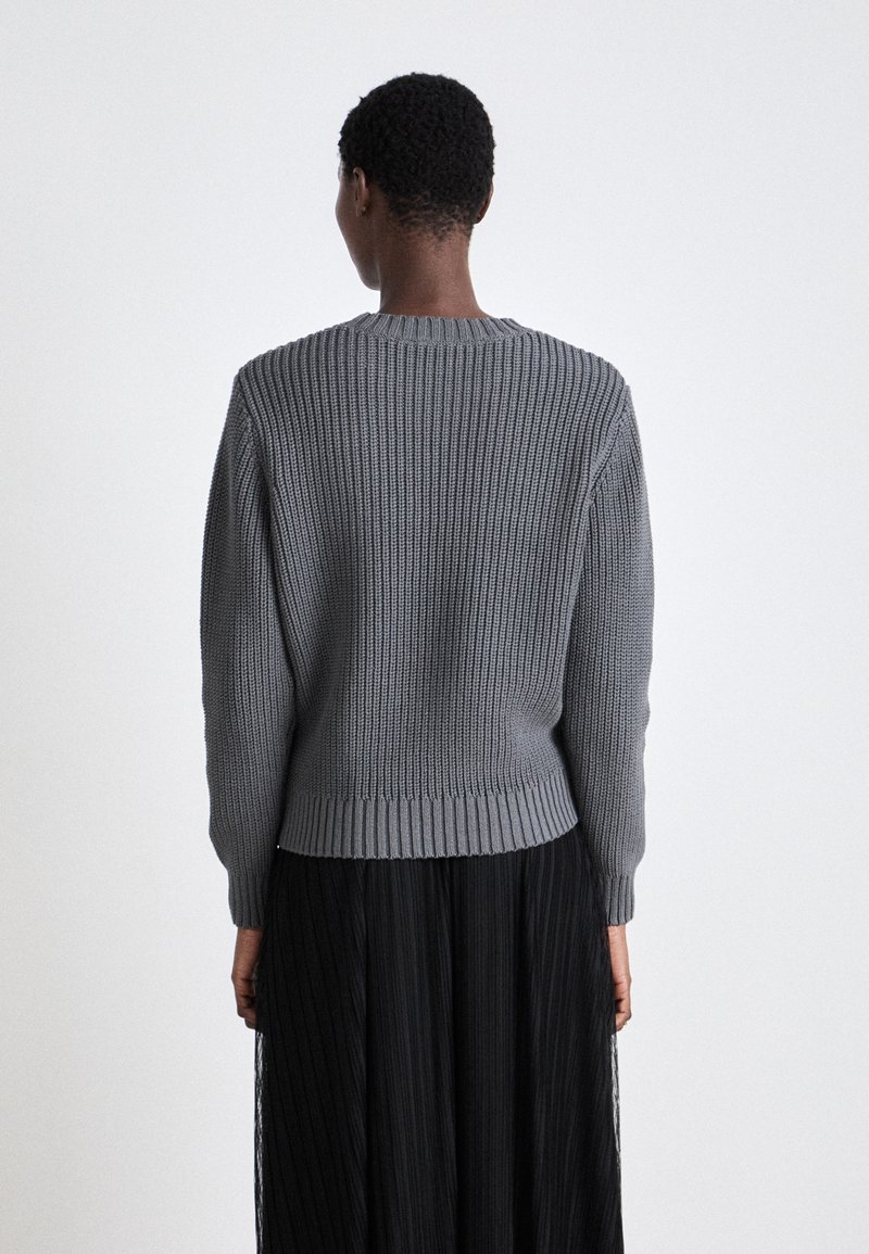 Grauer Strickpullover mit gerippter Textur, Rundhalsausschnitt und langen Ärmeln, kombiniert mit einem fließenden schwarzen Rock.