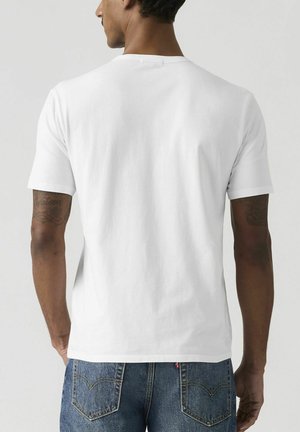 Mężczyzna o ciemnej karnacji i tatuażach, ubrany w prosty biały krótki rękaw koszulki i niebieskie dżinsy, stojący tyłem na jednolitym tle.