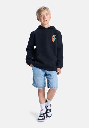 Sweat à capuche noir avec une poche kangourou, associé à un short en jean clair. Chaussures comprenant des baskets noires et blanches avec des accents rouges.