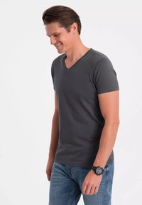 Šedé tričko s výstřihem do V a krátkými rukávy, vyrobené z hladké látky. Nosené s světle modrými džíny a černými hodinkami na levém zápěstí.