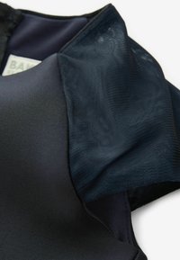 Schwarzes Kleidungsstück mit einem glatten, matten Stoff und durchsichtigen Mesh-Ärmeln. Die Texturen kontrastieren zwischen festen und durchsichtigen Materialien.