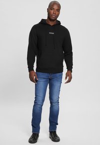 Black hooded sweatshirt with "GUESS" logo, front pocket, paired with blue jeans and black lace-up sneakers. Smooth fabric, casual fit.
