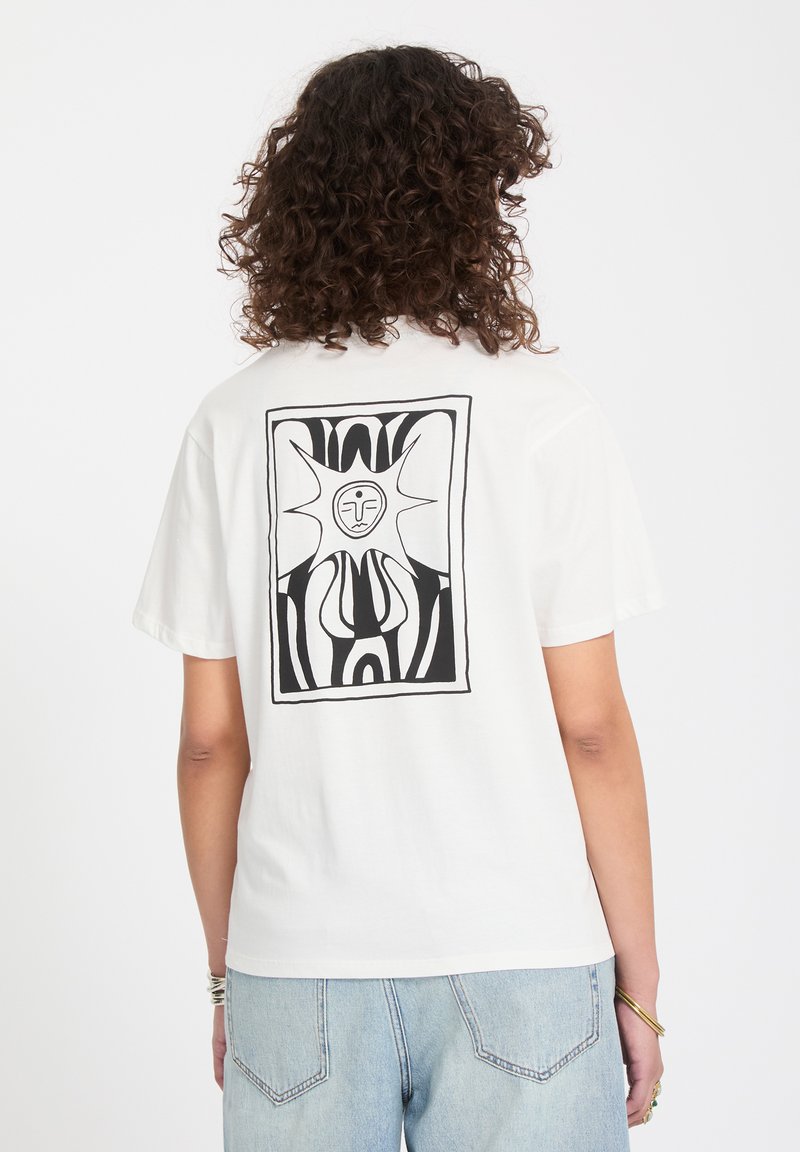 Personne aux cheveux bouclés portant un t-shirt blanc avec un motif abstrait noir représentant un soleil et un visage au dos, associé à un jean bleu clair.