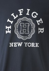 Tricou din bumbac bleumarin, cu un logo circular alb ce conține textul "HILFIGER" și "NEW YORK", iar în centru se află un "H" proeminent.