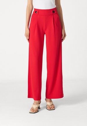 Red wide-leg trousers with pleats and black button accents at the waistband, made from a smooth fabric. Paired with beige heeled sandals.