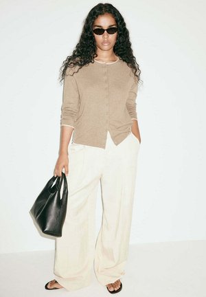 Femme aux longs cheveux bouclés portant des lunettes de soleil, un cardigan beige, un pantalon large blanc, des sandales noires et tenant un sac à main noir sur fond blanc.