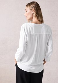 Camisa blanca de manga larga con un corte relajado, escote redondeado y textura suave; la parte posterior presenta un detalle plisado. Combinada con pantalones negros.