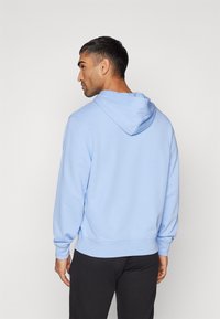 Helle blaue Kapuzenjacke mit Kordelzug, gerippten Bündchen und Saum. Weicher Stoff, bequemer Schnitt. Schwarze Hose ist an der Taille sichtbar.