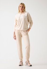 Béžová mikina s logem "GUESS" vyloženým kamínky, doplněná o sladěné zúžené kalhoty. Hladká tkanina a volný design. Tenisky s akcenty.