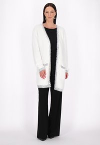 Weißer, flauschiger Cardigan mit schwarzem Besatz, offener Vorderseite, langen Ärmeln und Taschen, getragen über einem schwarzen Outfit mit weit geschnittenen Hosen.