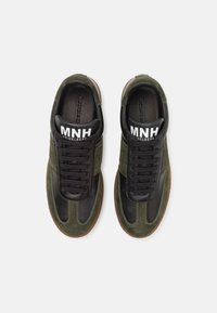 Sneaker in pelle nera e suede verde con suola in gomma marrone, dotati di lacci tondi e un grande logo bianco sulla linguetta.