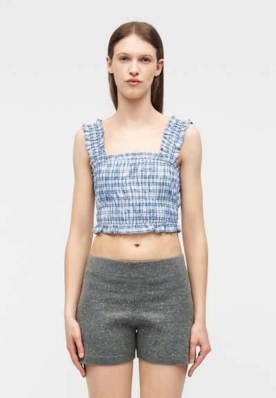 Jeune femme portant un crop top sans manches à carreaux bleus et blancs et un short en maille grise avec un motif à pois subtil.