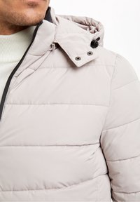 Světle šedá prošívaná bunda s kapucí, s horizontálním prošíváním, předním zipem a patenty na límci. Měkká textura.