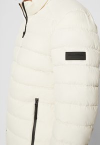 Giacca puffer bianca con design a cuciture orizzontali, zip nera e una patch del logo nera sulla manica sinistra. Finitura liscia e colletto alto.