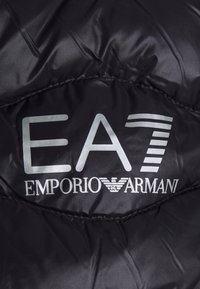 Czarna, pikowana tkanina z wyraźnym srebrnym logo "EA7 EMPORIO ARMANI" oraz dekoracyjnym szyciem i gładką fakturą.