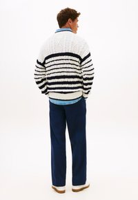 Ριγέ πουλόβερ σε λευκό και ναυτικό μπλε, με πλεκτή υφή καλωδίου, συνδυασμένο με σκούρο μπλε παντελόνι και λευκά αθλητικά παπούτσια. Εμφανίζεται η πίσω όψη.