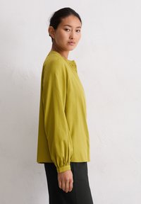 Langsleeve shirt in lichtgroen, gemaakt van zachte stof. Heeft een ronde halslijn en geplooide mouwen, gecombineerd met zwarte broek.