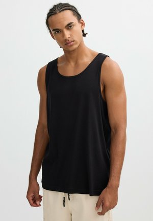 Joven con cabello trenzado, vestido con una camiseta sin mangas negra y pantalones beige, posando frente a un fondo claro y liso.