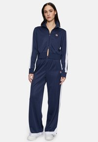 Marineblauwe trainingsoutfit met streepdessin; korte ritssluitingen jas en high-waisted broek; witte zijstrepen; stof heeft een gladde textuur.