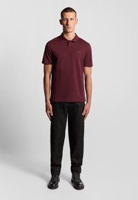 Bordeaux polo-shirt med krave, korte ærmer og et diskret logo. Sammen med sorte bukser og solide sorte sko med mønstrede snørebånd.