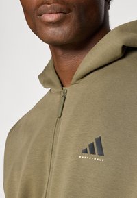 Olive green zip-up hoodie made of soft fabric, featuring a black adidas logo and "BASKETBALL" text. Smooth texture with a relaxed fit.
