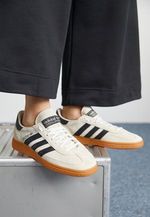Beige suède sneakers met een zwart drie-streep ontwerp en witte veters, voorzien van een getextureerde rubberen zool en het 'adidas' logo.