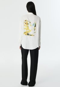 Chemise blanche texturée présentant un imprimé floral jaune et bleu au dos. Assortie à un pantalon large noir. Tissu doux, coupe allongée.