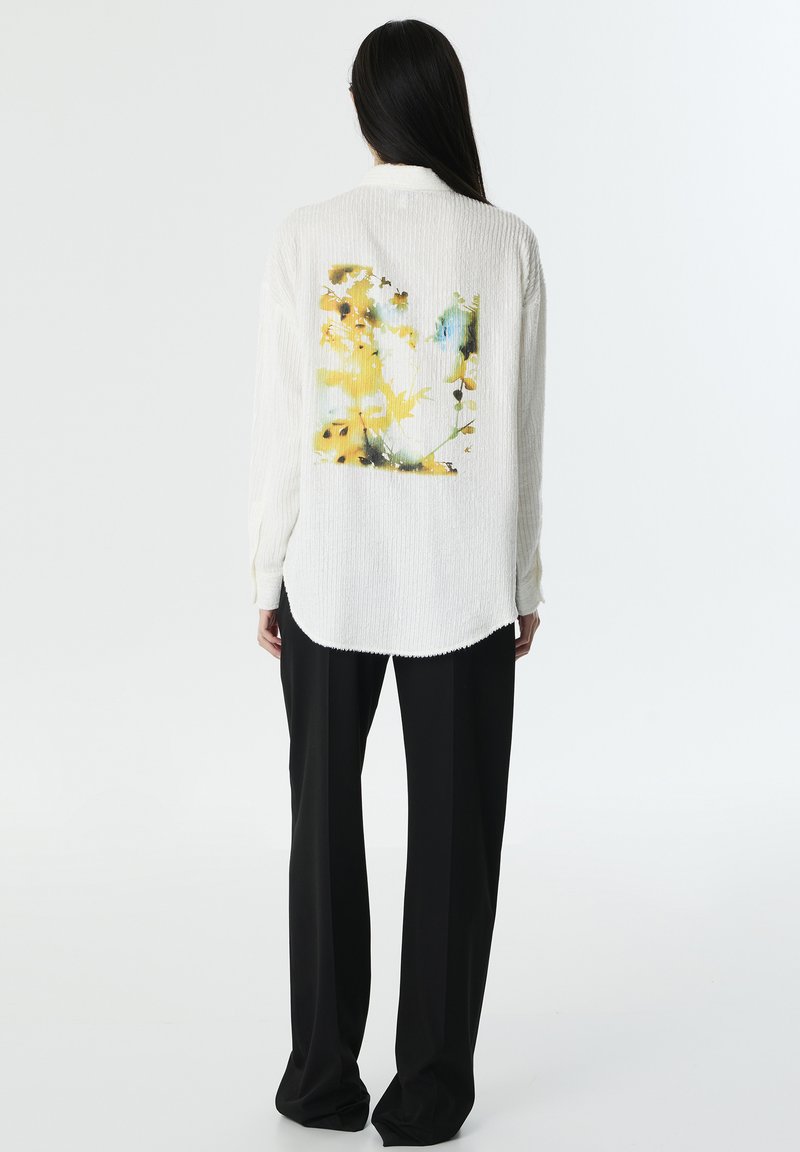 Chemise blanche texturée présentant un imprimé floral jaune et bleu au dos. Assortie à un pantalon large noir. Tissu doux, coupe allongée.