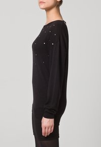 Schwarzer Strickpullover mit langen Ärmeln, rundem Halsausschnitt und vorne mit kleinen goldenen Nieten verziert. Weiche Textur und entspannte Passform.