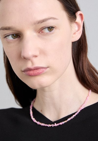 Ροζ κολιέ από χάντρες με ανώμαλα σχήματα και λεία υφή, φορεμένο πάνω από ένα μαύρο ένδυμα, αναδεικνύοντας τις πολύχρωμες λεπτομέρειές του.